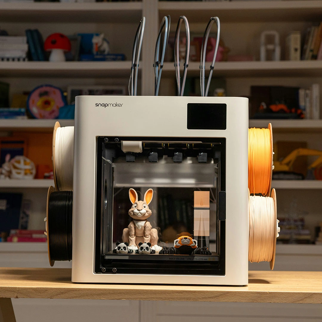 Profesjonalna drukarka 3D Snapmaker z wieloma szpulami filamentu drukująca kolorowe figurki na biurku w domowym warsztacie.